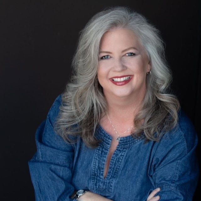 A smiling upper-torso image of Jenny Booth with long grey-ombre wavy hair and a denim tunic.