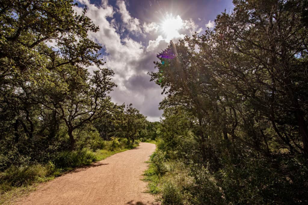 The photo depicts a gravel trail with green trees and shrubs on either side. The sky is visible with the sun breaking through the clouds.