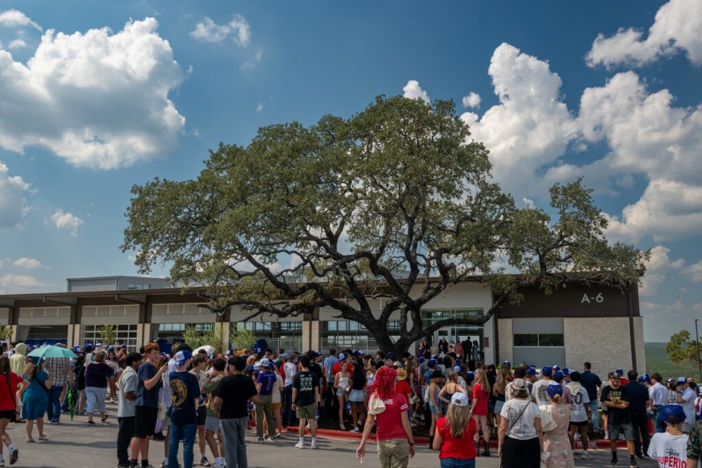 A crowd mingling outside of the aquatic center on opening day near an oak tree with cumulous clouds.